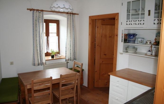 Kleine Küche mit Holzmöbeln, Esstisch, Stühlen und einem Fenster mit Vorhängen.