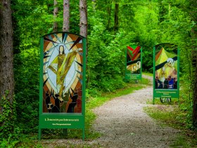 Weg durch einen Wald, links und rechts neben dem Weg stehen gro&szlig;e Tafeln aus Glas, die die Stationen des Kreuzweges darstellen.