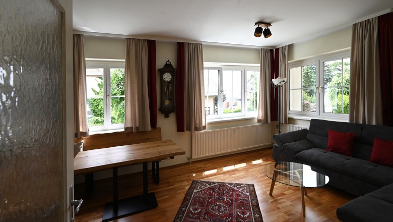 Ein Wohnzimmer mit Holzboden, grauem Sofa, Glastisch, Wanduhr und Teppich. Gro&szlig;e Fenster mit Vorh&auml;ngen lassen viel Licht herein.