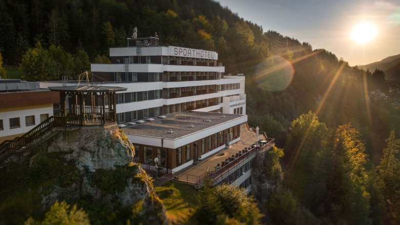 Ein Hotel auf einem H&uuml;gel bei Sonnenuntergang, umgeben von Wald.