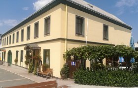 Gelbes Gasthaus mit grünen Fensterrahmen, Gastgarten und Pflanzen vor dem Eingang.