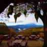 Terrasse mit Holztischen und B&auml;nken, umgeben von B&auml;umen, mit Blick auf bewaldete Berge und Wolken am Horizont.