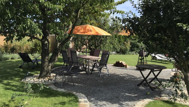 Garten mit Sitzbereich unter B&auml;umen, Tisch und St&uuml;hlen, orangefarbener Sonnenschirm.