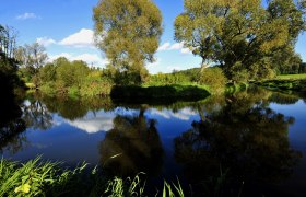 Ein ruhiger Fluss im Naturpark Dobersberg mit B&auml;umen und blauem Himmel im Hintergrund.
