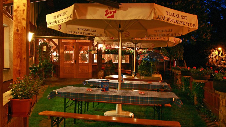 Ein gem&uuml;tlicher Biergarten bei Nacht mit Holztischen, blau karierten Tischdecken und gro&szlig;en Sonnenschirmen der Marke Stiegl.