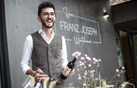 Mann mit Brille und Weste h&auml;lt Weinflasche vor Restaurant 'Franz Joseph'.