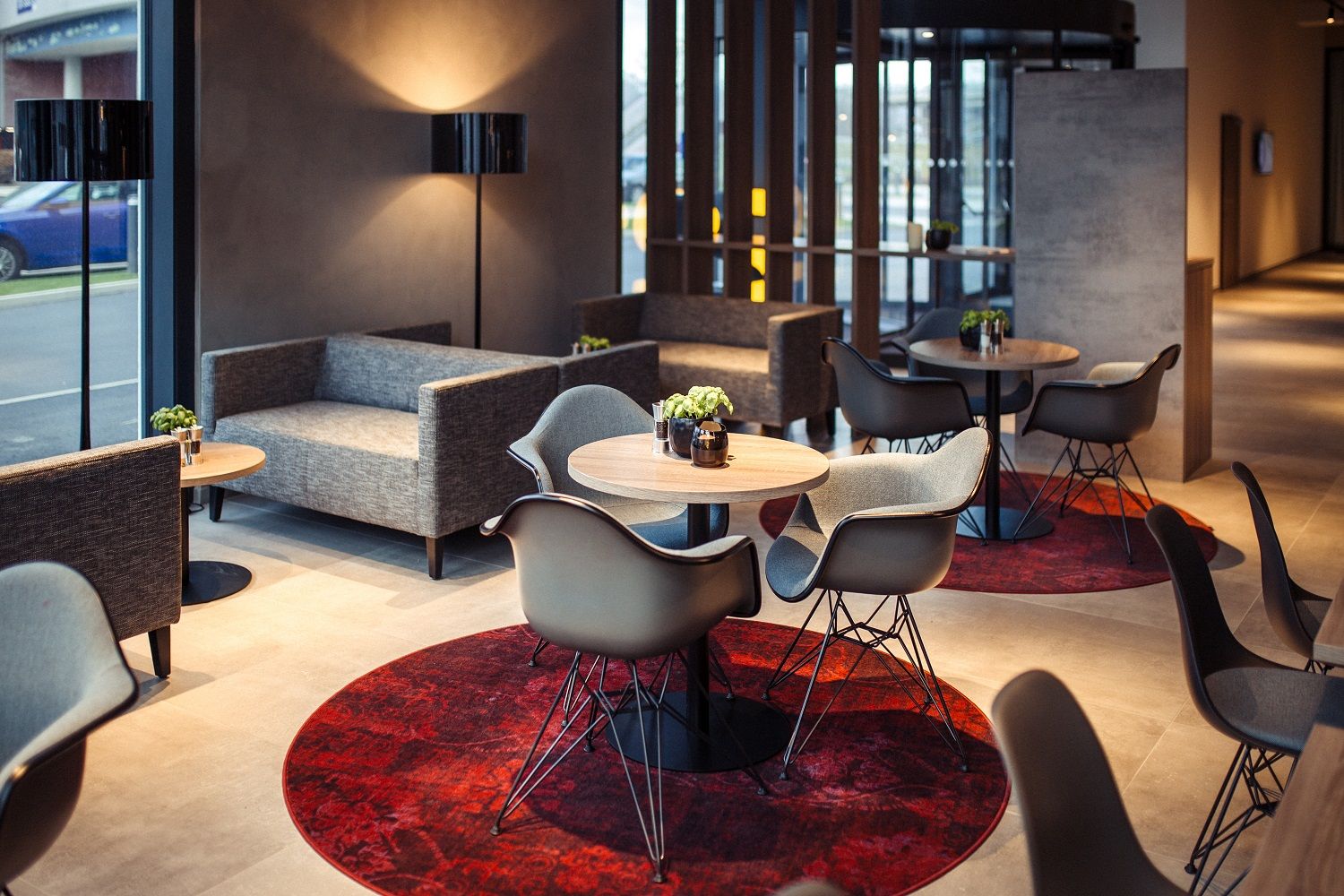 Moderne Hotellobby mit grauen Sofas, runden Holztischen und roten Teppichen.
