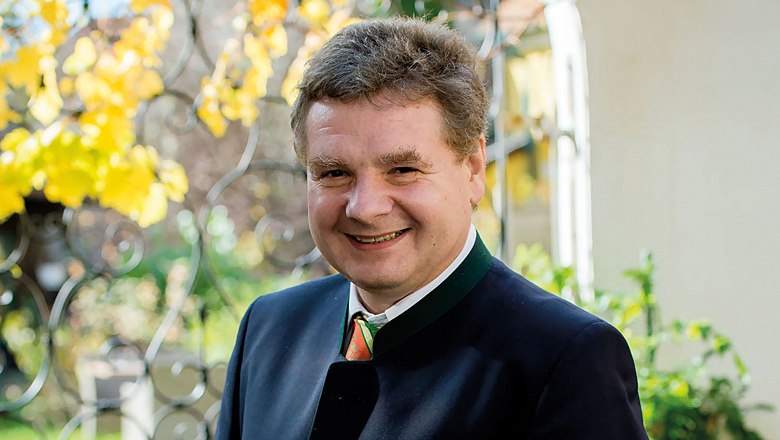 Ein l&auml;chelnder Mann in traditioneller Kleidung steht im Freien vor einem schmiedeeisernen Zaun mit gelben Bl&auml;ttern im Hintergrund.