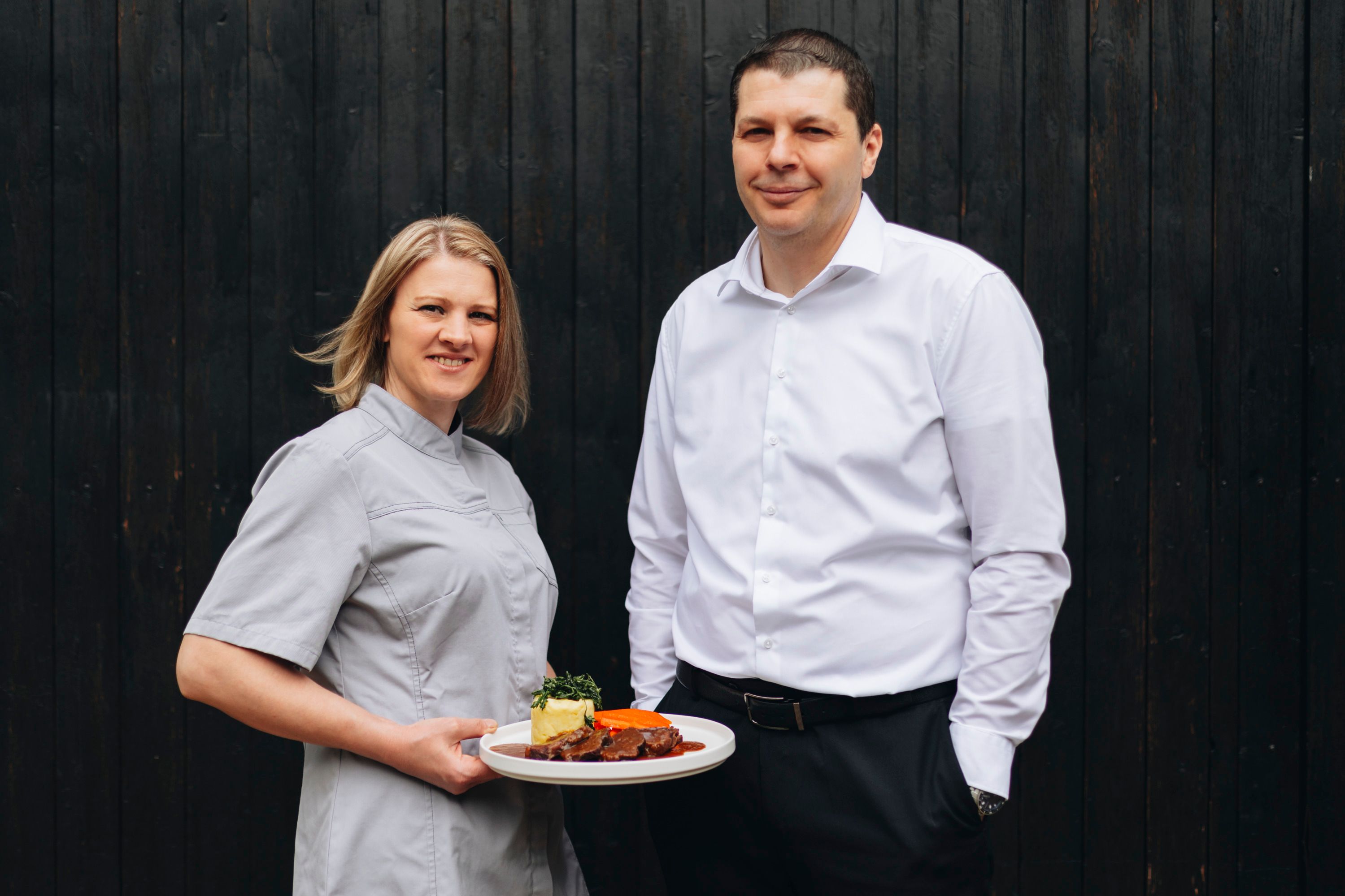 Zwei Personen, eine Frau in Kochkleidung mit einem Teller, vor dunklem Hintergrund.