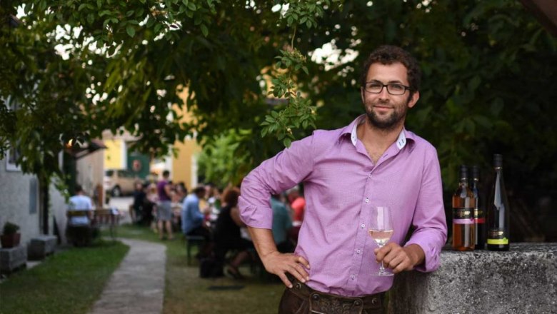 Mann in lila Hemd mit Weinglas, steht draußen neben Flaschen.