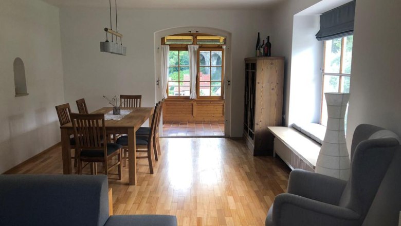 Ein Wohnzimmer mit Holzboden, Esstisch, Stühlen, Sessel und Fenster mit Blick nach draußen.