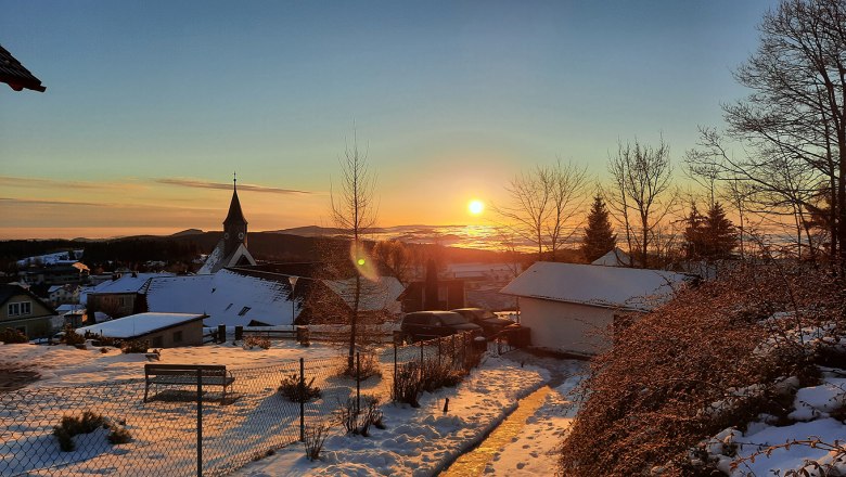 Winterlicher Sonnenaufgang &uuml;ber einer verschneiten Landschaft mit Kirche und H&auml;usern.