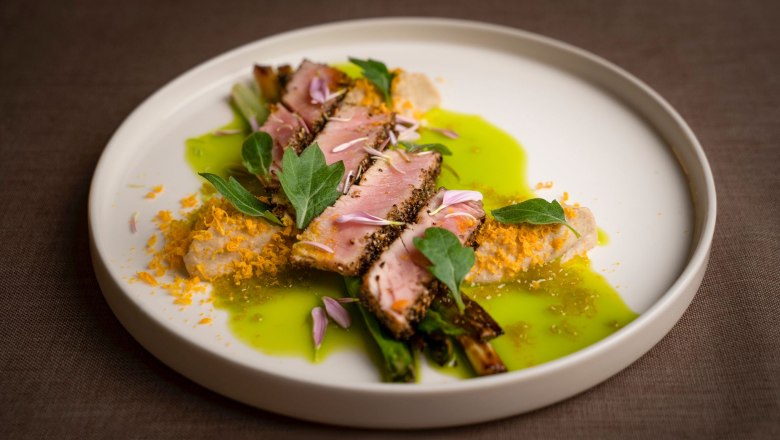 Ein kunstvoll angerichtetes Gericht mit Fleisch, gr&uuml;ner Sauce und essbaren Bl&uuml;ten auf einem wei&szlig;en Teller.