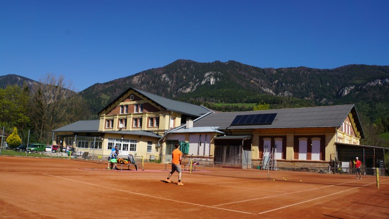 Tennisplatz vor einem Gebäude mit Bergen im Hintergrund.