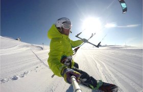 Person beim Snowkiten auf einer verschneiten Piste mit blauem Himmel und Sonnenschein.
