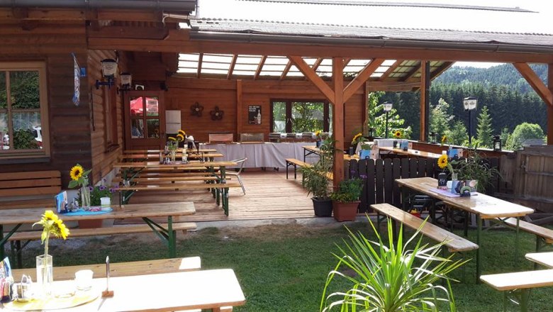 Ein gem&uuml;tlicher Biergarten mit Holztischen und B&auml;nken, dekoriert mit Sonnenblumen, umgeben von gr&uuml;ner Landschaft.