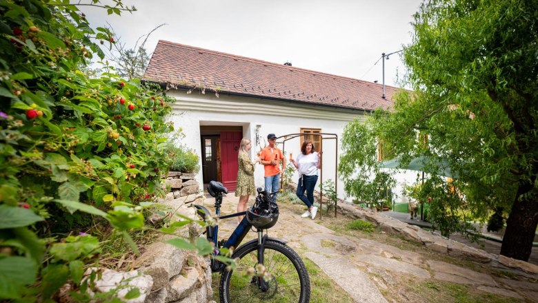 Drei Personen stehen vor einem weißen Haus mit rotem Dach, ein Fahrrad im Vordergrund.