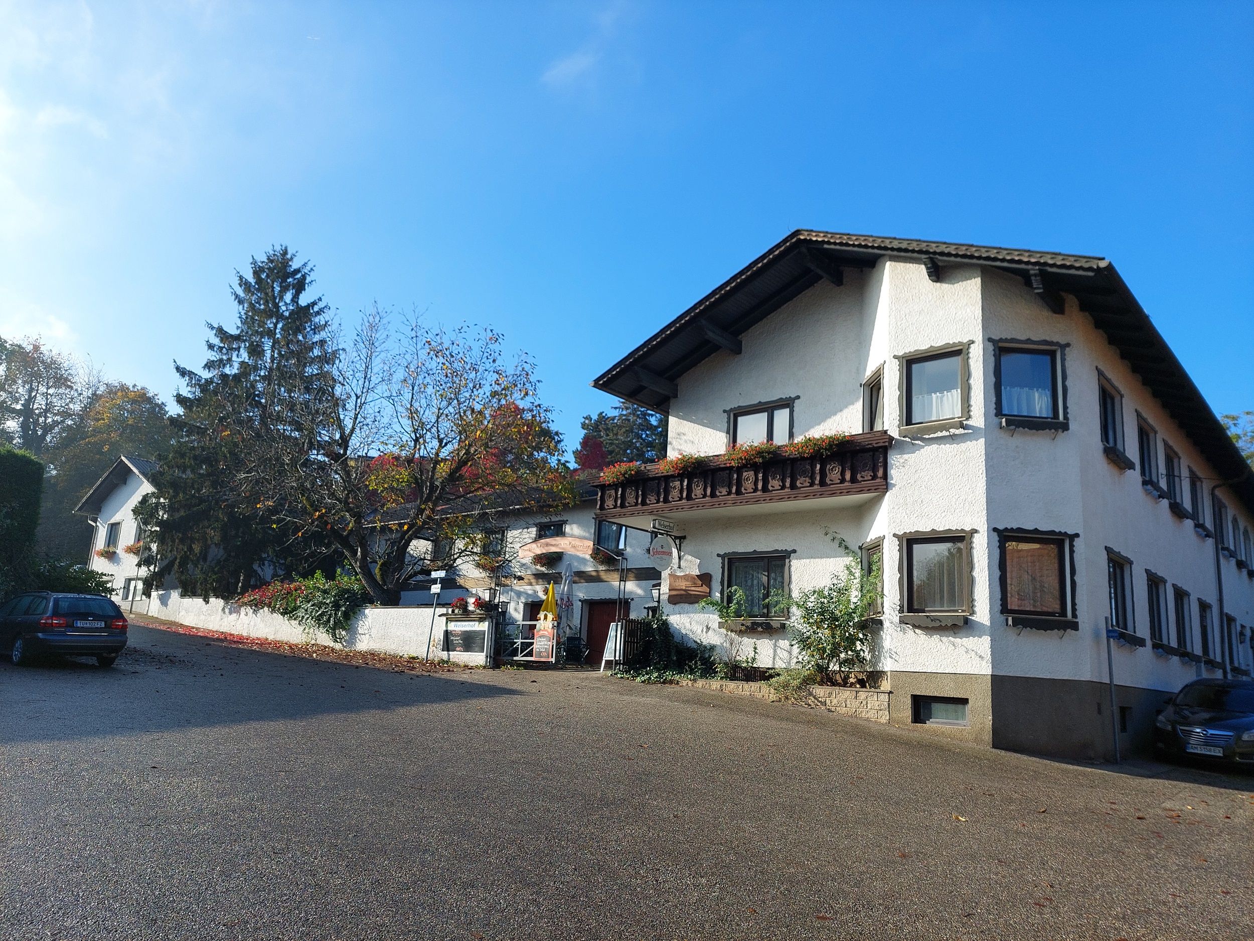 Außenansicht eines traditionellen Gebäudes mit weißen Wänden und Holzverzierungen, umgeben von Bäumen und einem blauen Himmel.