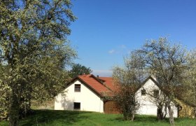 Ein weißes Haus mit rotem Dach steht auf einer grünen Wiese, umgeben von blühenden Bäumen unter einem klaren blauen Himmel.