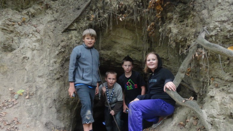 Vier Kinder stehen am Eingang einer kleinen H&ouml;hle im Wald.