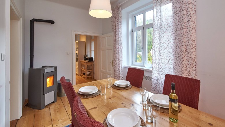 Esszimmer mit Holztisch, roten St&uuml;hlen, gedecktem Tisch und Kaminofen.