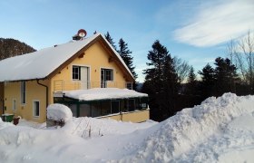 Ein gelbes Haus mit verschneitem Dach und Balkon, umgeben von Bäumen und Schnee.