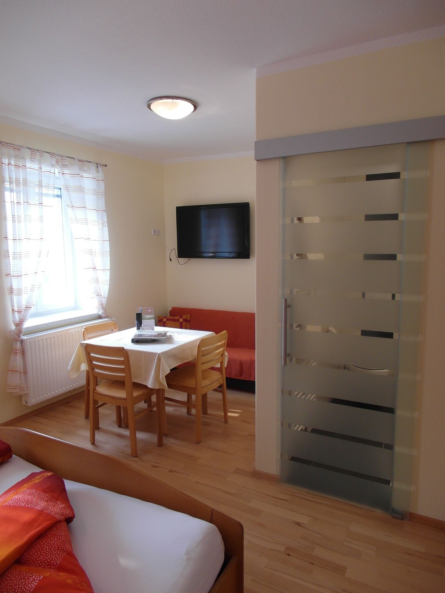 Helles Zimmer mit Essecke, Tisch, Stühlen, Bett und Flachbildfernseher.