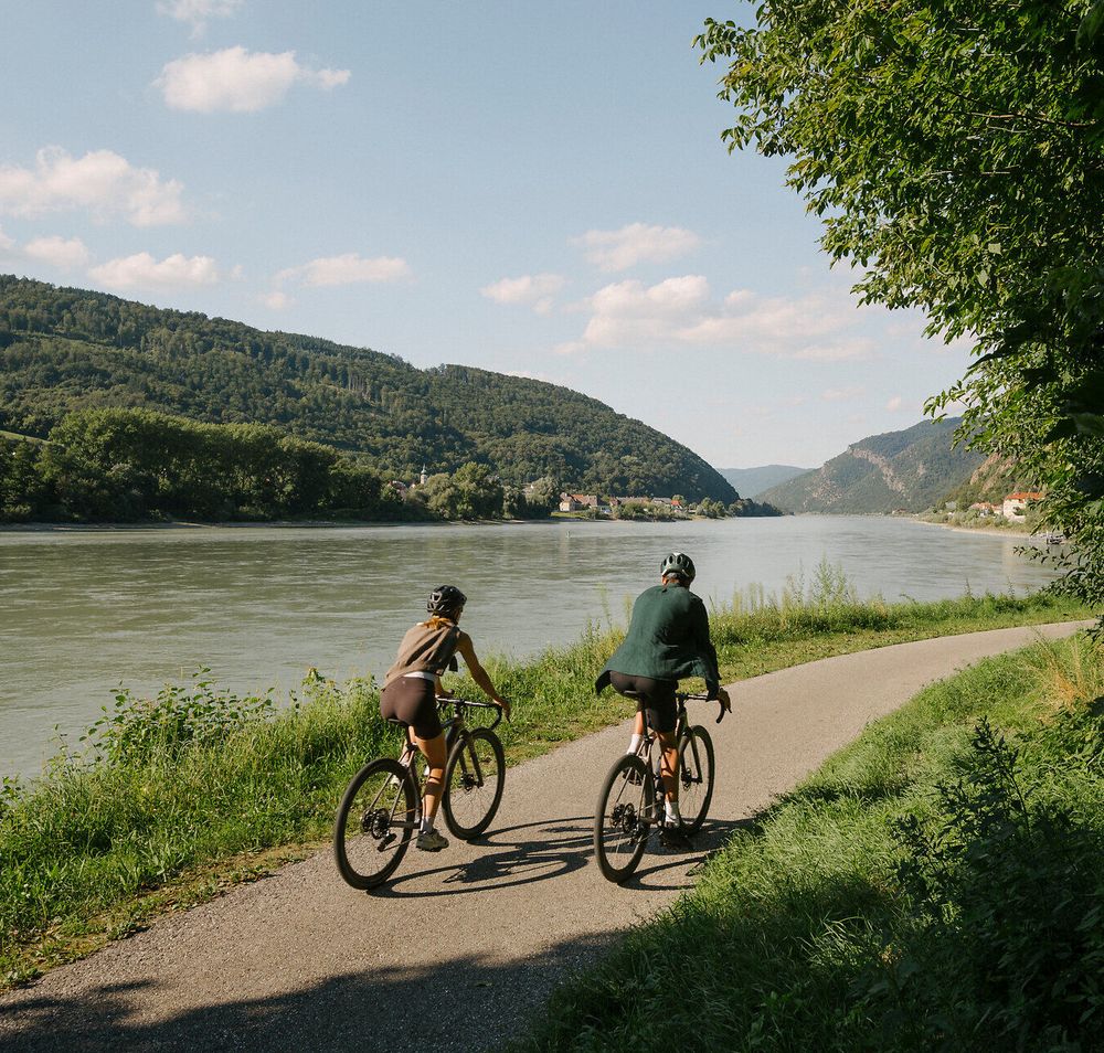 Zwei Radfahrende auf einem asphaltierten Uferweg entlang der Donau; gegenüber liegen bewaldete Hügel, rechts spenden Bäume Schatten.