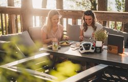 Zwei Radfahrerinnen sitzen auf einer Terrasse und essen an einem Tisch; darauf stehen Getränke, Speisen und ein Fahrradhelm.