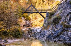 Die goldenen Herbstfarben spiegeln sich im klaren Wasser des Baches wider, während die sanften Geräusche des plätschernden Wassers eine friedliche Atmosphäre schaffen. Eine malerische Holzbrücke verbindet die beiden Ufer und lädt dazu ein, die atemberaubende Natur des Naturparks Ötscher Tormäuer zu erkunden.
