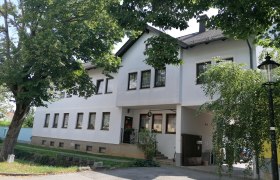 Außenansicht eines weißen Gebäudes mit vielen Fenstern, umgeben von Bäumen.