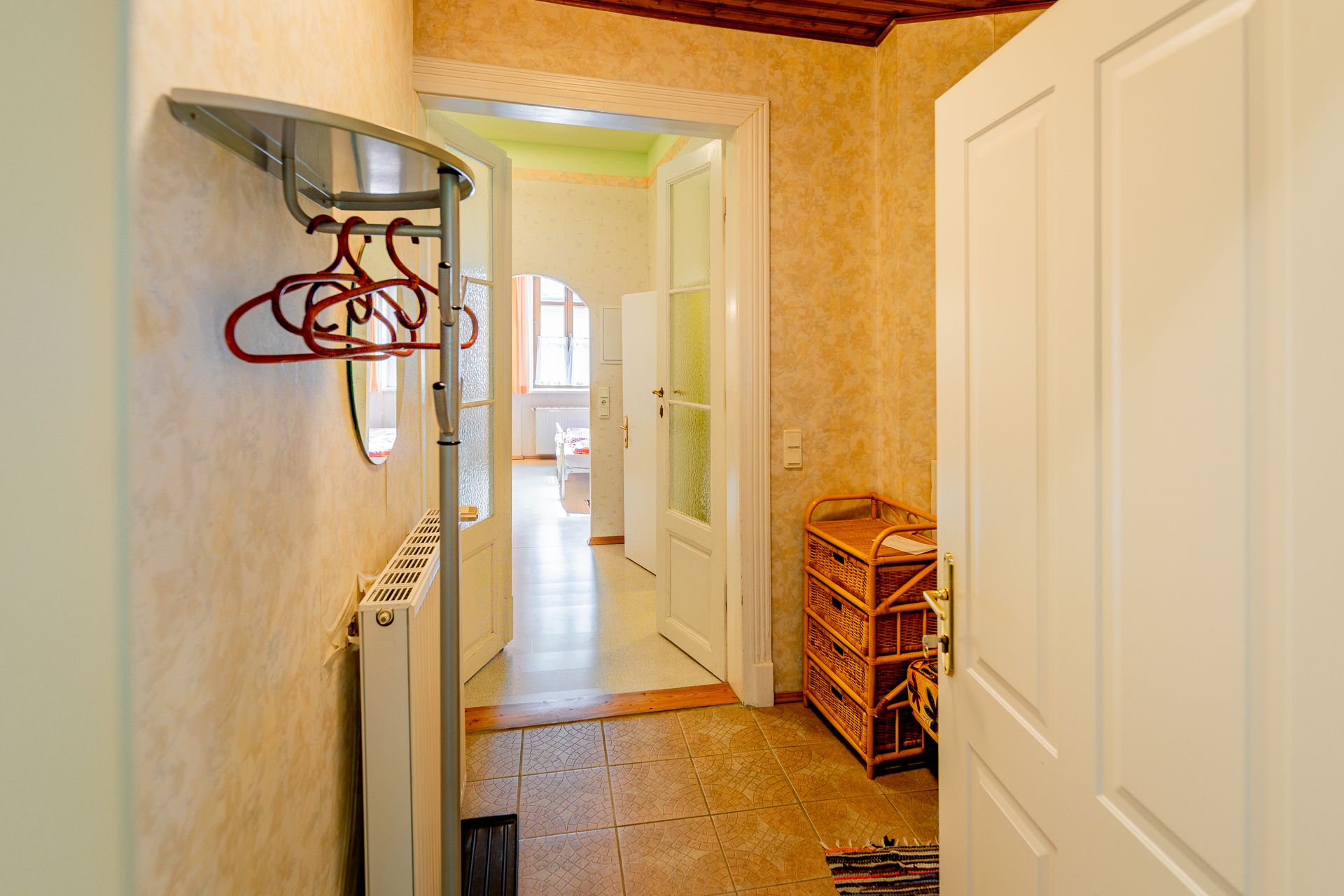 Ein Vorraum mit Kleiderhaken, einem Heizkörper und einem Korbmöbel. Eine Tür führt in einen hellen Raum mit Fenstern.