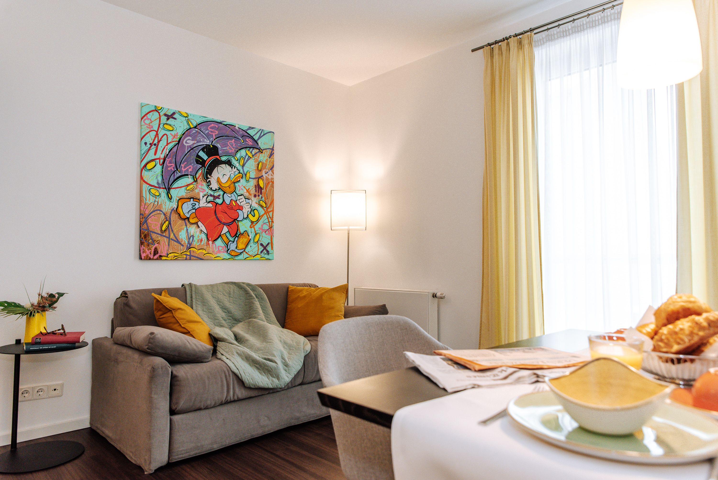 Wohnzimmer mit grauem Sofa, bunter Comic-Kunst an der Wand, gelben Vorhängen und Frühstückstisch.