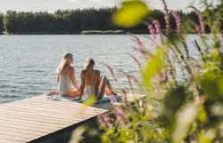 Zwei Personen in Badebekleidung sitzen auf Handtüchern auf einem Holzsteg am Viehofner See, im Hintergrund ist ein bewaldetes Ufer zu sehen.