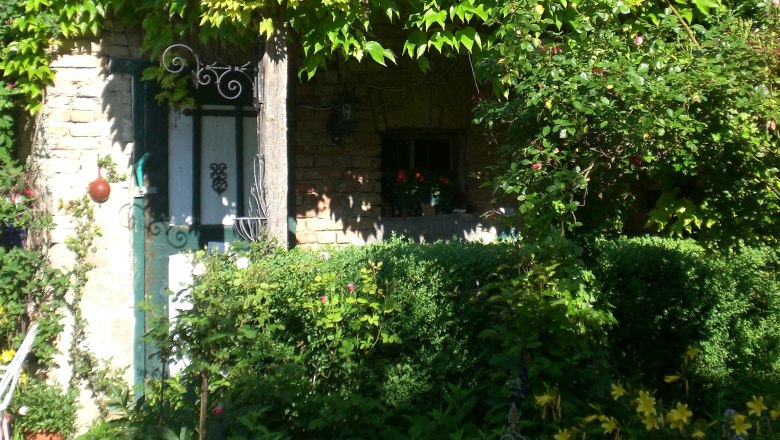 Ein idyllischer Landhausgarten mit &uuml;ppigem Gr&uuml;n und Blumen vor einem alten Steinhaus.