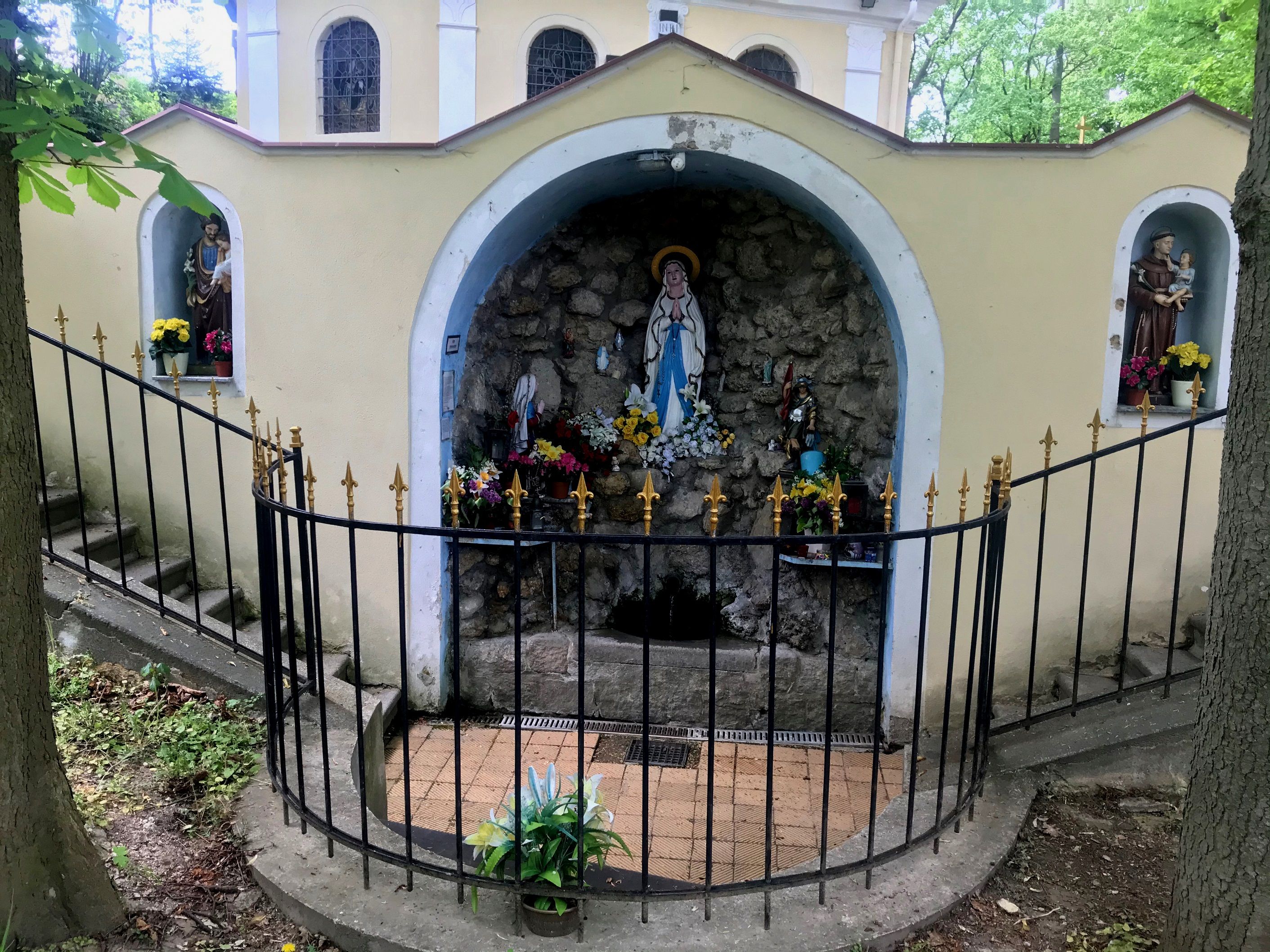 Eine Grotte mit religiösen Statuen und Blumen, umgeben von einem schmiedeeisernen Zaun.