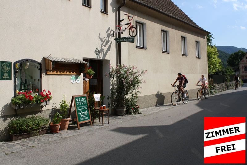 Außenansicht eines Hauses mit Fahrraddekoration und Weinausschank. Zwei Radfahrer fahren vorbei. Schild mit 'Zimmer frei'.