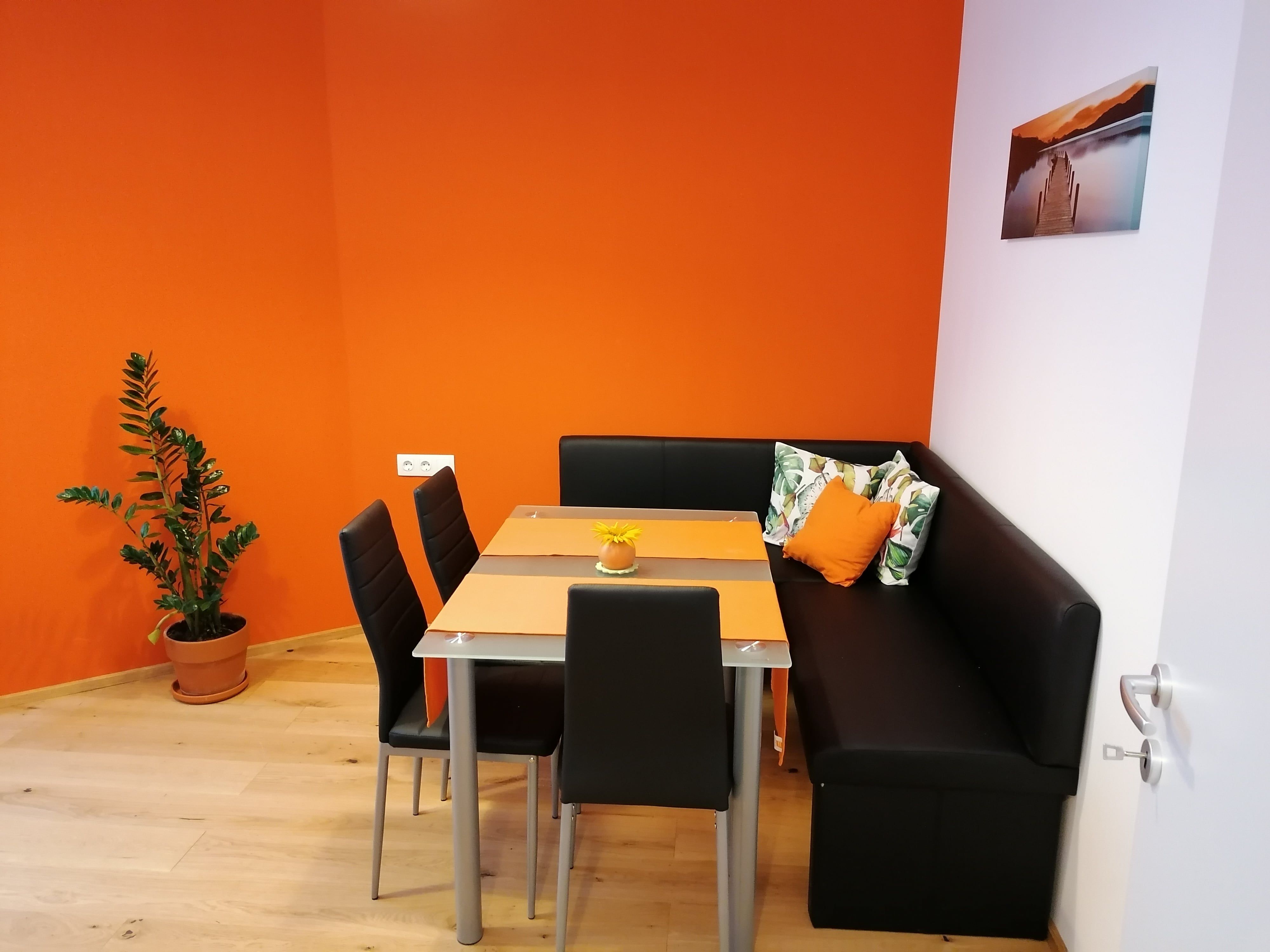 Ein moderner Frühstücksraum mit oranger Wand, schwarzer Eckbank, Tisch und Stühlen. Eine Pflanze steht in der Ecke, ein Bild hängt an der Wand.