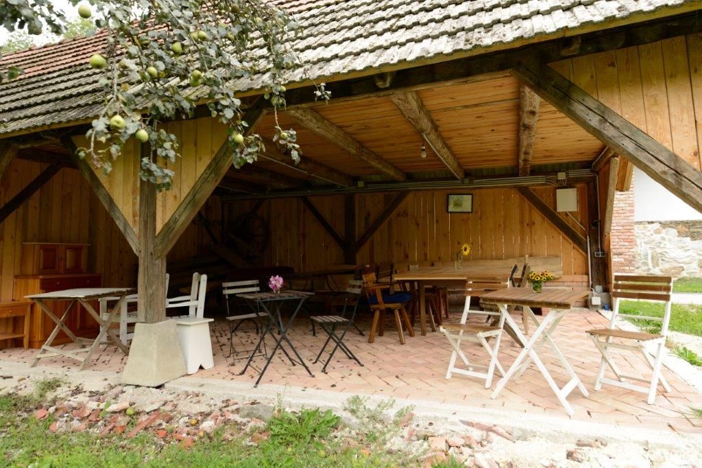 Überdachte Terrasse mit Holztischen und Stühlen, umgeben von Holzwänden und einem Apfelbaum im Vordergrund.