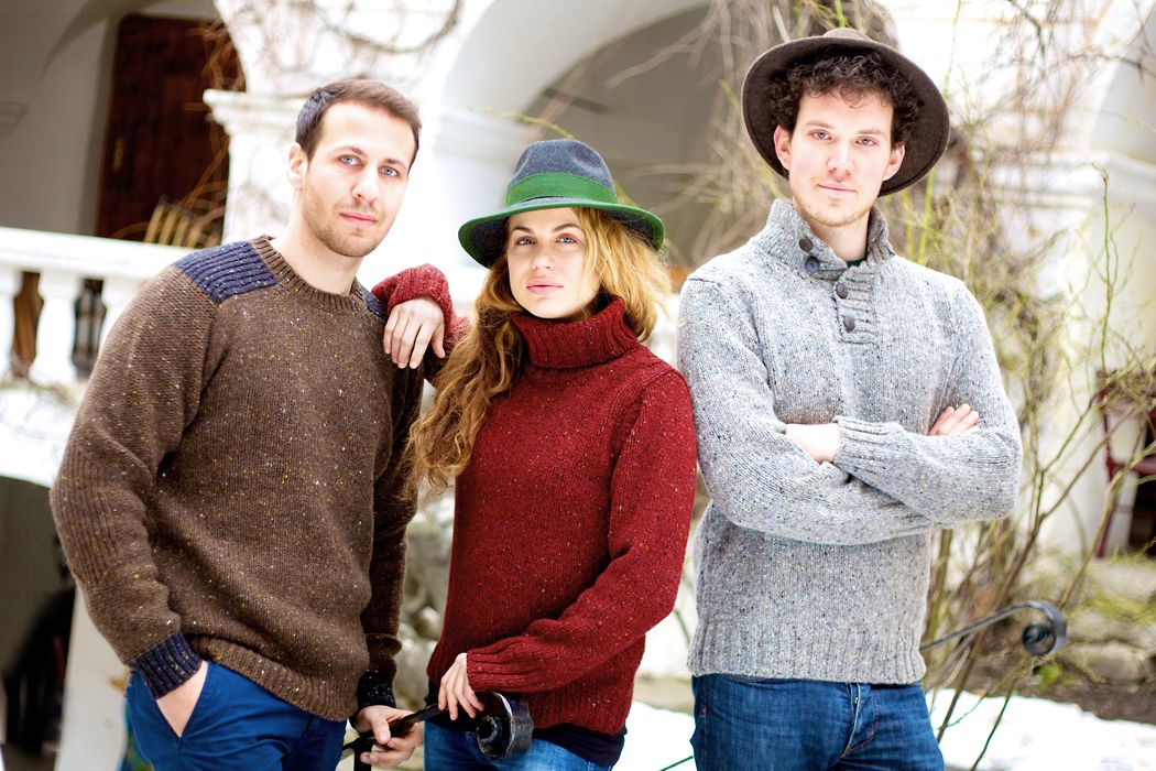 Drei Personen in Wollpullovern und Hüten stehen draußen vor einem Gebäude.