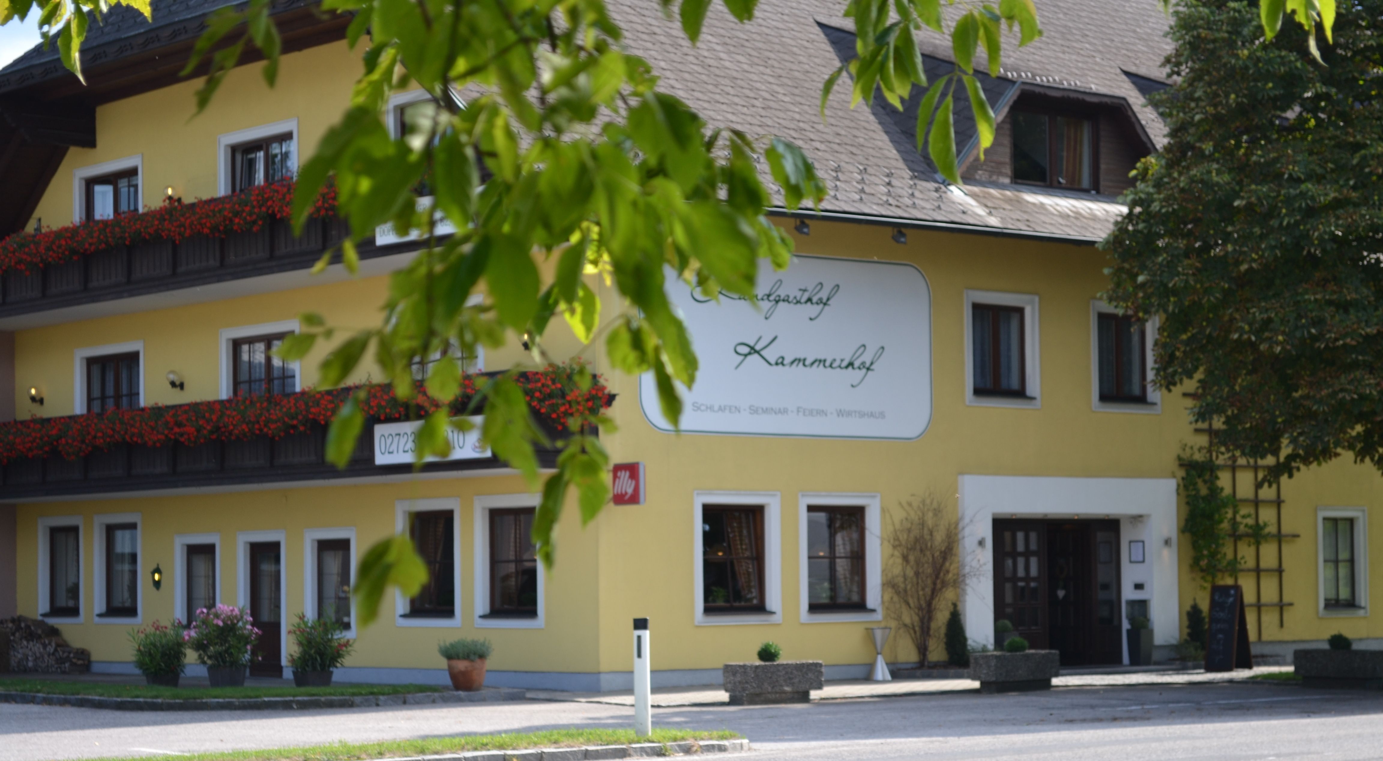 Gelbes Gebäude mit Schild 'Landgasthof Kammerhof', Blumenkästen und Bäumen im Vordergrund.