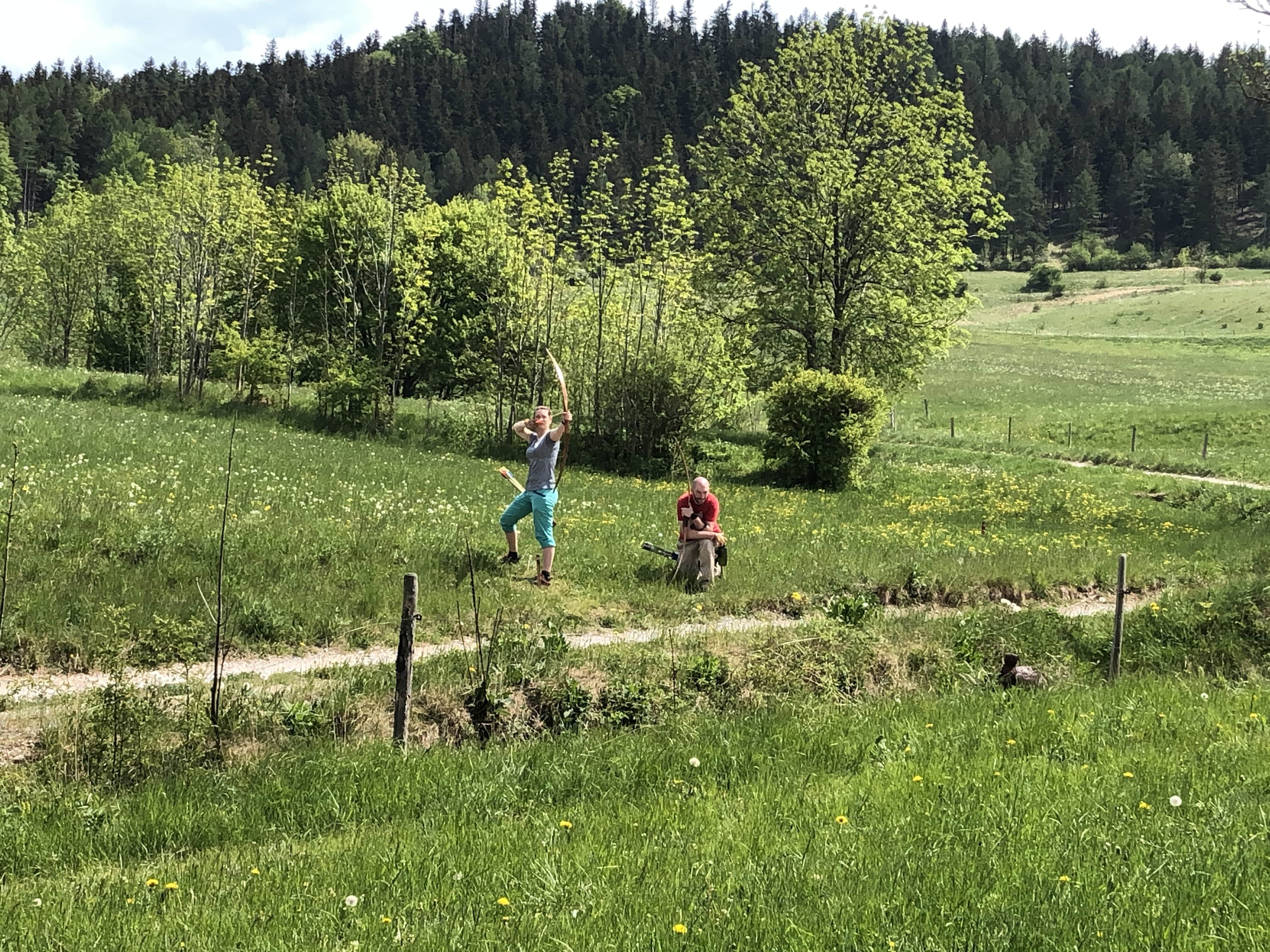 Zwei Personen auf einer Wiese, eine schießt mit einem Bogen, die andere sitzt. Im Hintergrund Bäume und Hügel.
