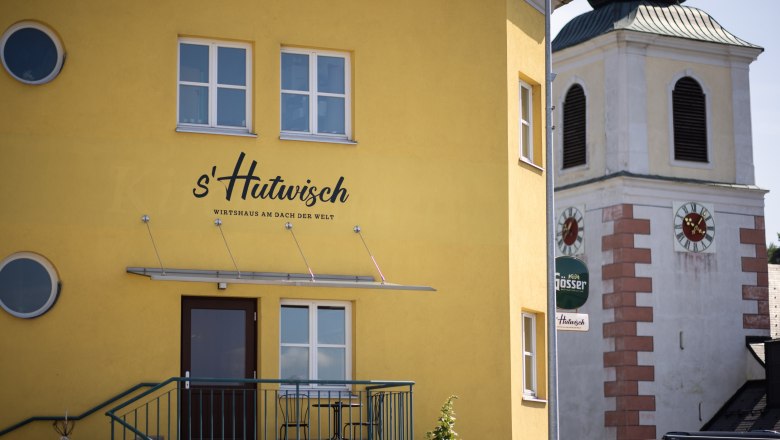 Gelbes Geb&auml;ude mit der Aufschrift 's'Hutwisch' neben einem Kirchturm mit Uhr.
