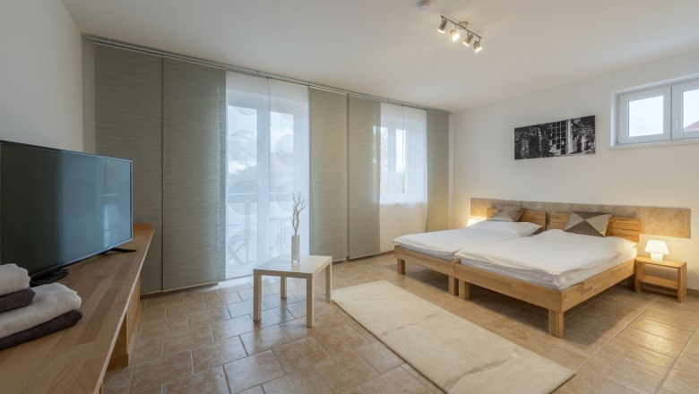 Modernes Schlafzimmer mit Doppelbett, Fernseher, Tisch und großen Fenstern mit Vorhängen.