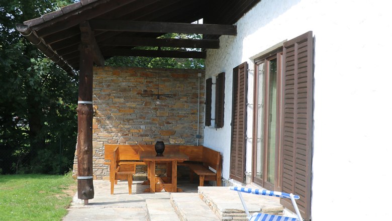 Terrasse mit Liege und Holzm&ouml;beln neben einem Haus mit Schindeldach.