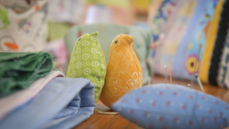 Zwei Stoffv&ouml;gel, einer gr&uuml;n und einer orange, auf einem Tisch mit Stoffen und N&auml;hutensilien.