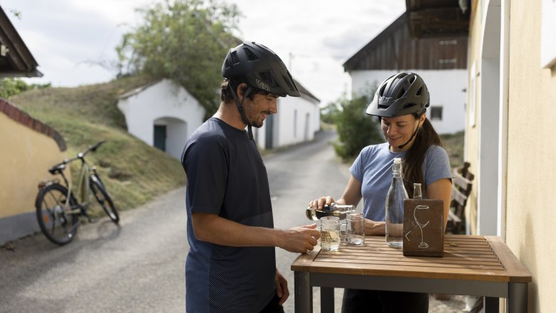 Zwei Radfahrer mit Helmen stehen an einem Tisch in einer Kellergasse und schenken sich Getr&auml;nke ein.