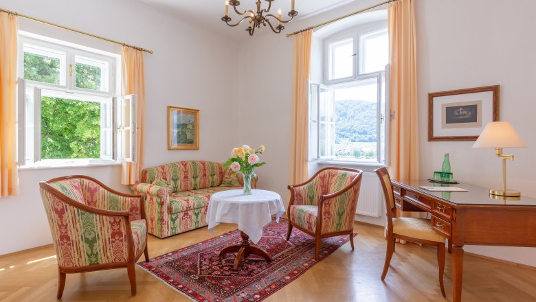 Elegantes Wohnzimmer mit antiken M&ouml;beln, zwei Fenstern, einem Tisch mit Blumen, einem Schreibtisch und einem Teppich auf Holzboden.
