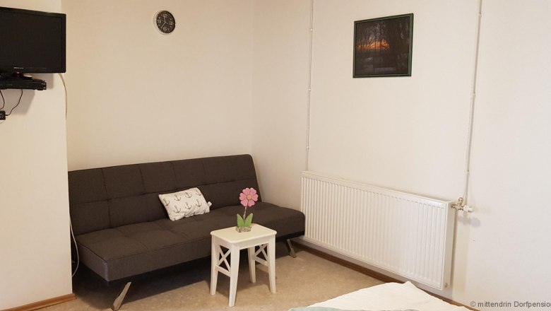 Fernseher an der Wand, daneben ein graues Sitzsofa und einem kleinem Tisch.