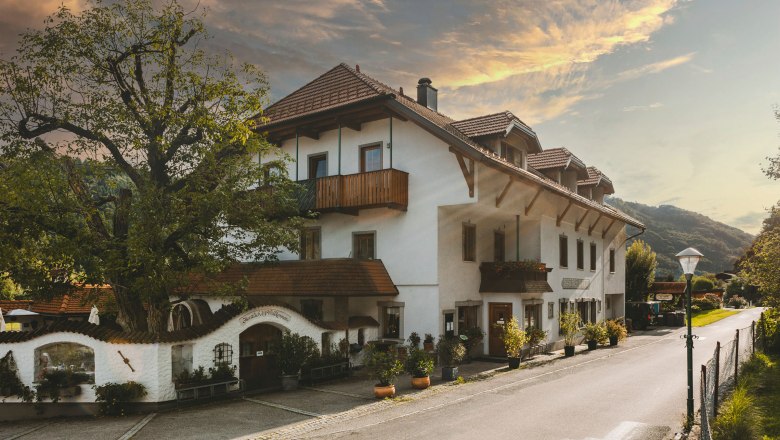 Ein traditionelles Wirtshaus mit wei&szlig;en W&auml;nden und roten Ziegeld&auml;chern, umgeben von B&auml;umen und einer ruhigen Stra&szlig;e.